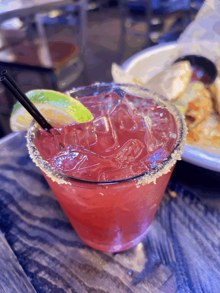 Spicy Blackberry Margarita- A deep-pink cocktail in a short glass with ice, a salted rim, a lime, and two stirring straws.